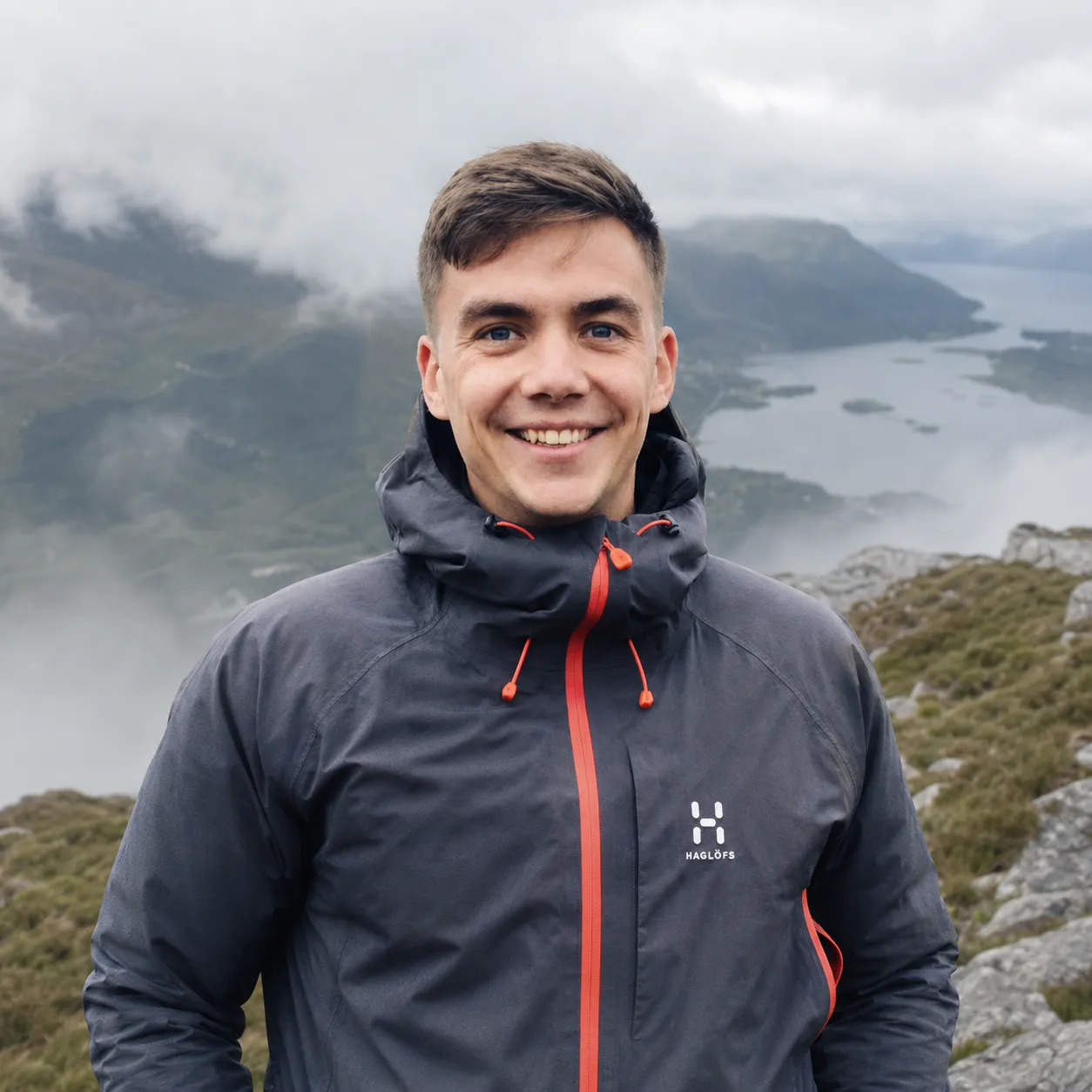 Louis Newman on a mountain summit above a fjord, smiling, wearing a dark shell jacket.